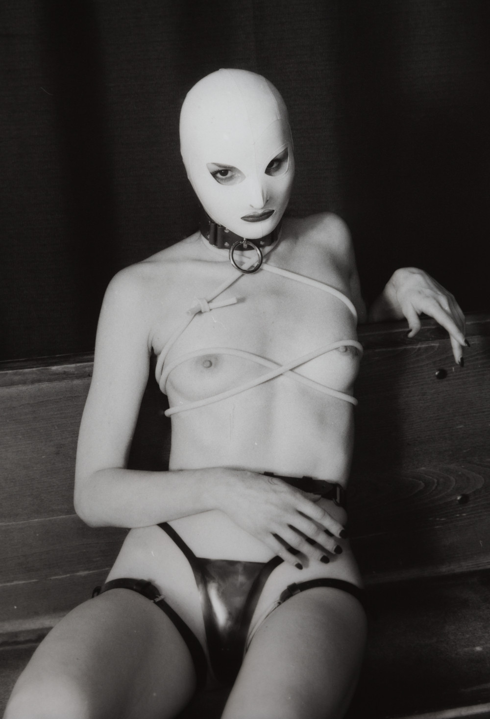 Magdalena Nurse IV by Johnny Welch. Photograph of a semi-nude woman wearing a white latex hood, choker, latex underwear, harness and tourniquet
