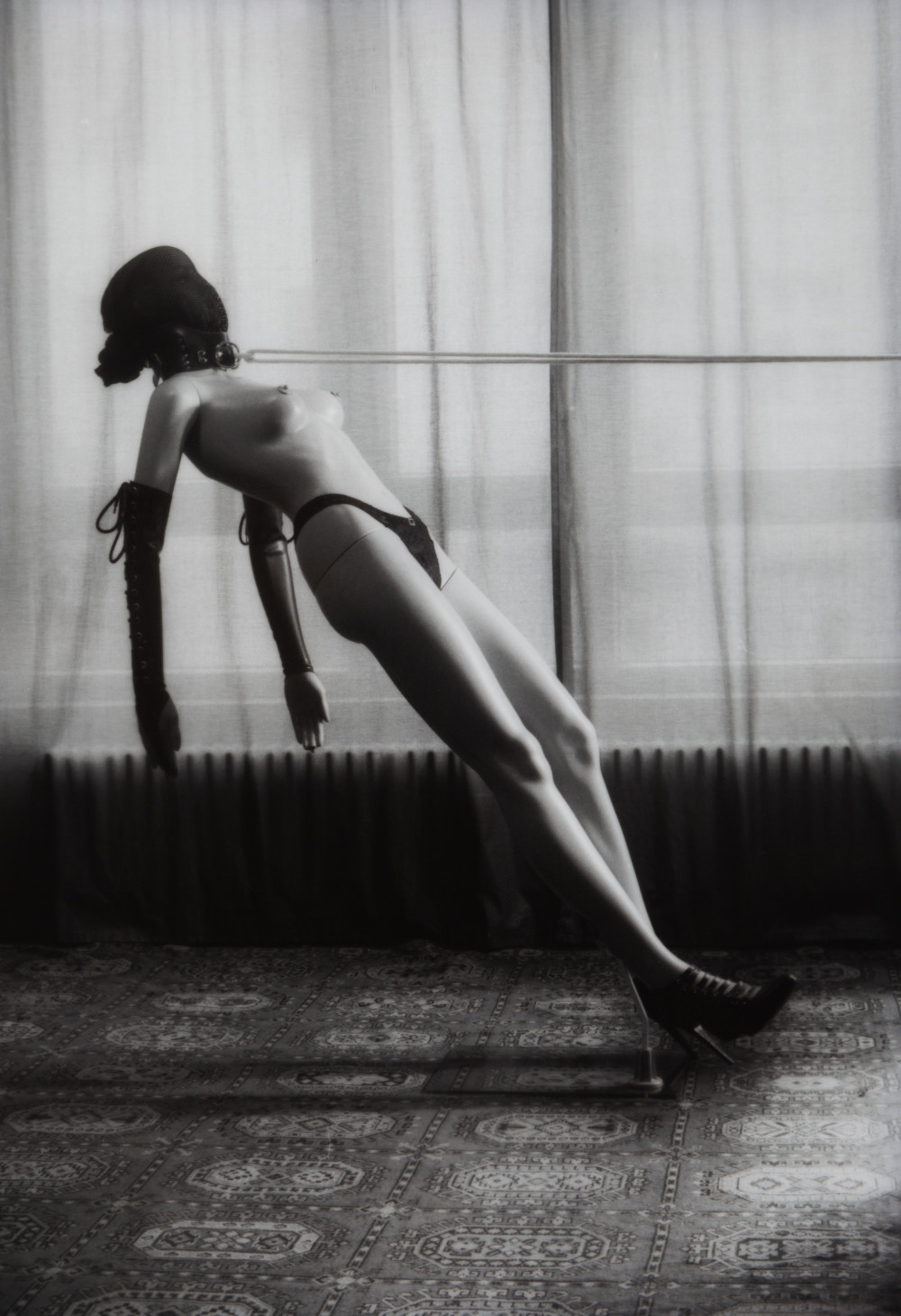 Mannequin Rope Bunny I by Johnny Welch. Photograph of a semi nude mannequin wearing black underwear, high heels, latex arm warmers, and a black mesh hood, tied with rope from a leather collar