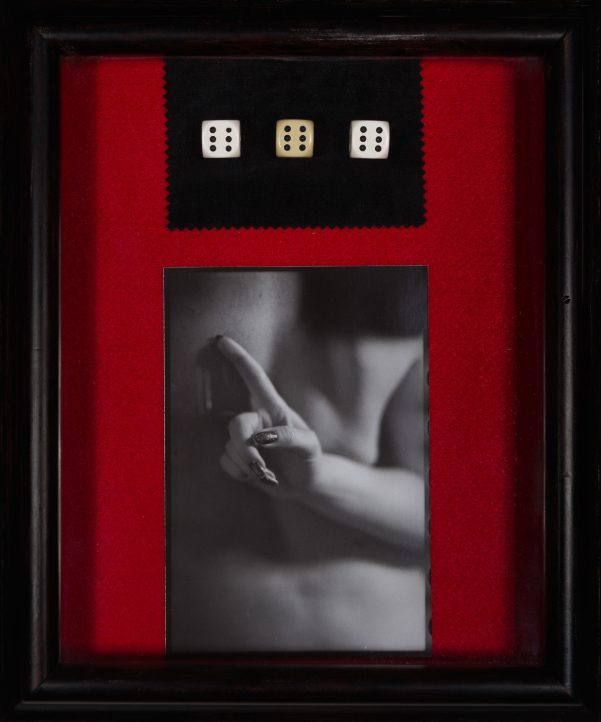 Olya 666 by Johnny Welch. Assemblage consisting of a photograph of a nude woman's hand behind her back, three dice showing the 6-face upright, a black cloth, mounted on red felt inside a black frame