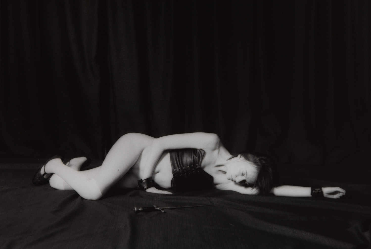 Kaya Athame III by Johnny Welch. A semi-nude woman lying on the ground next to a sword wearing a leather harness, cuffs and stockings