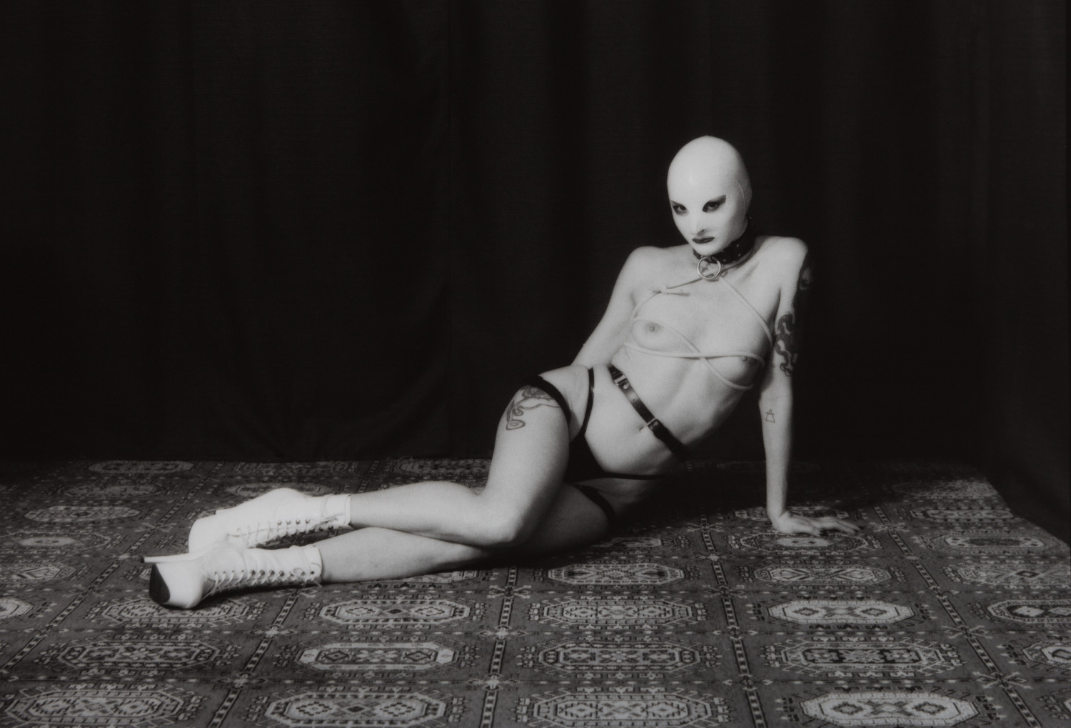 Magdalena Nurse VII by Johnny Welch. Photograph of a semi-nude woman wearing a white latex hood, choker, latex underwear, harness, boots and tourniquet